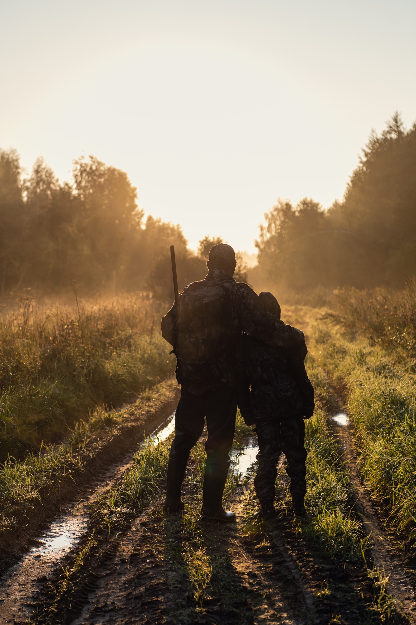 Hunters with hunting equipment going away through rural field towards forest at sunset during